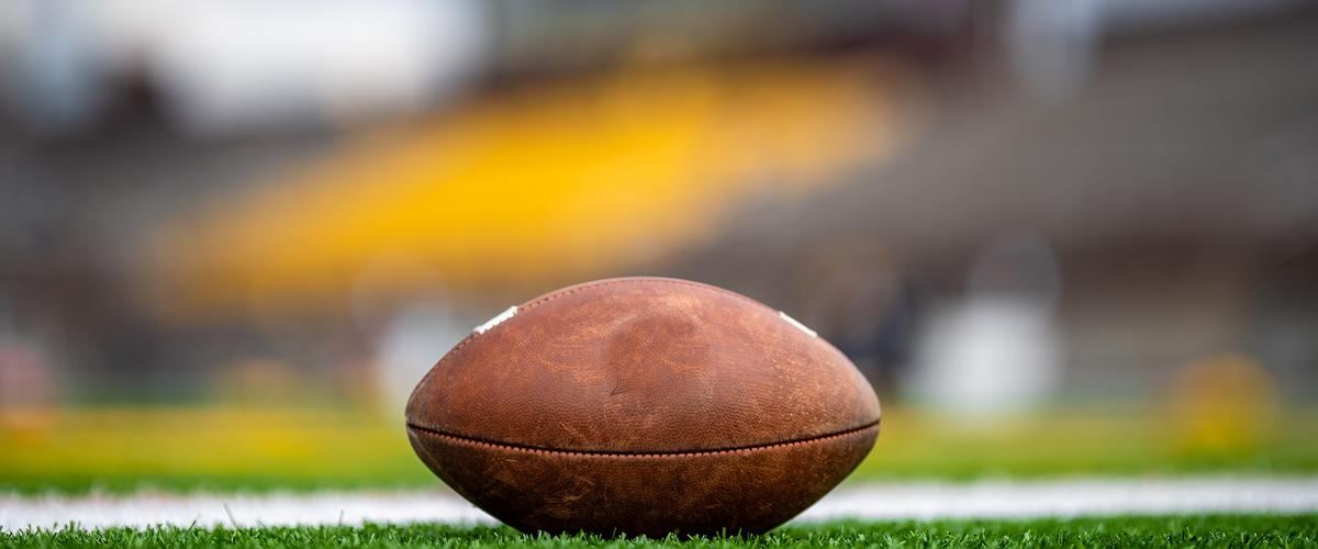 A brown leather football resting on green turf by a white yard line, with blurred yellow stadium seats behind it