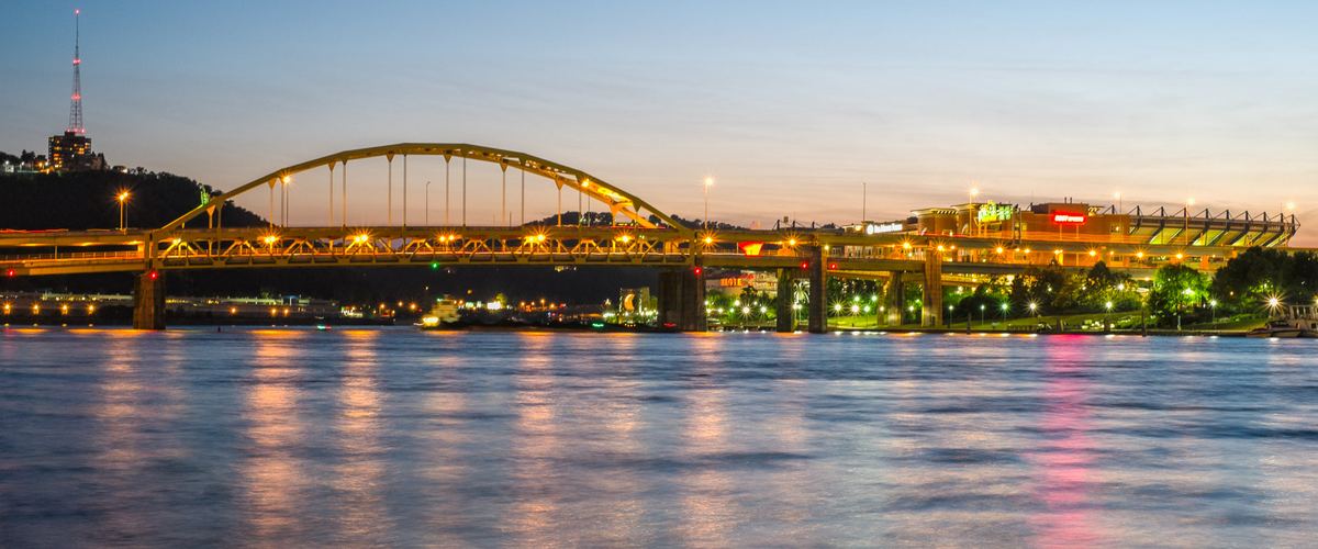 The lit-up yellow arch of the Fort Duquesne Bridge over the Allegheny River at dusk, with the North Shore stadium glowing in the distance