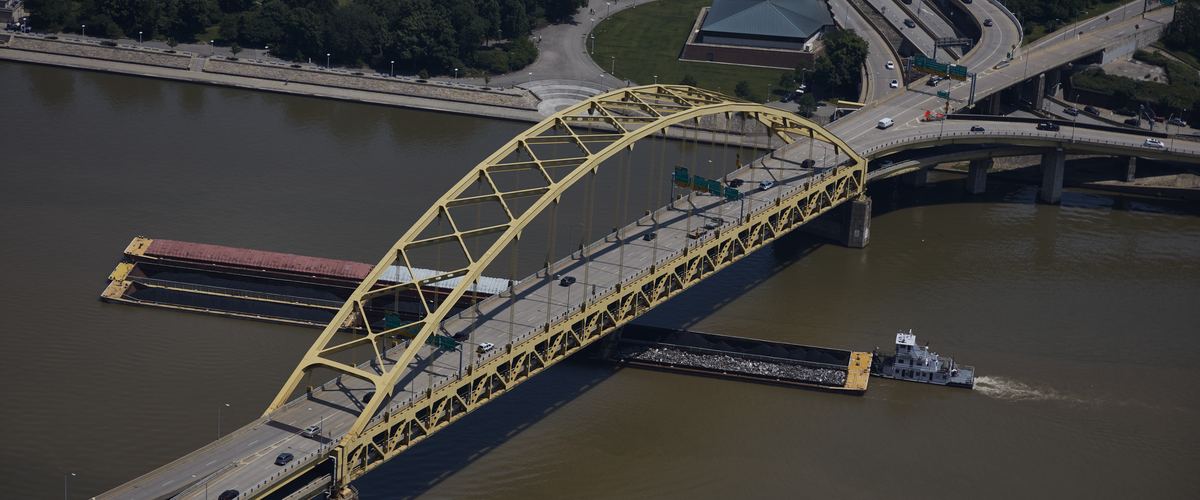 Aerial view of a tugboat pushing barges under the yellow arch of the Fort Pitt Bridge on the Monongahela River
