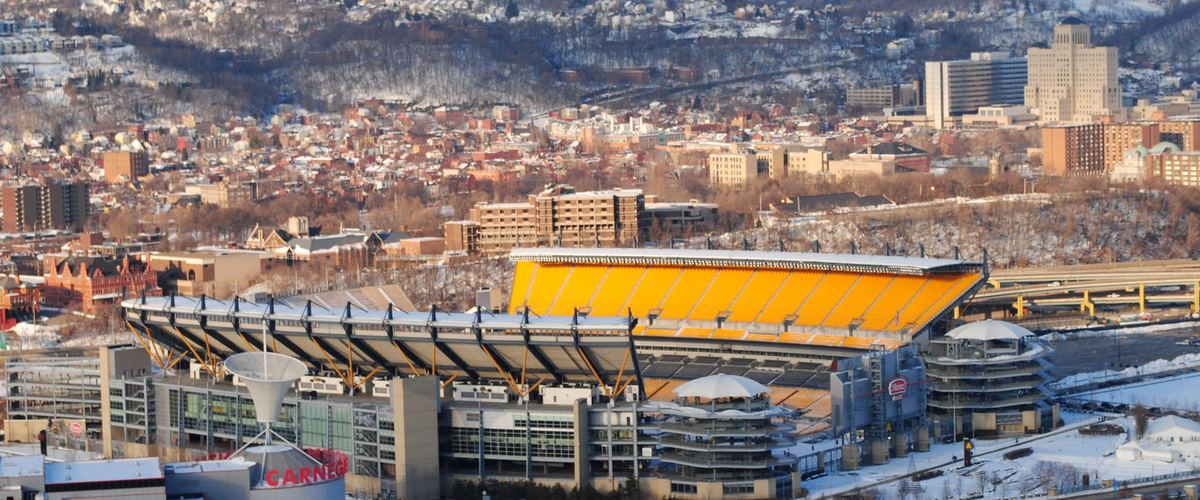 The North Shore football stadium with snow on the ground, seen from above across the river, with hillside neighborhoods behind it