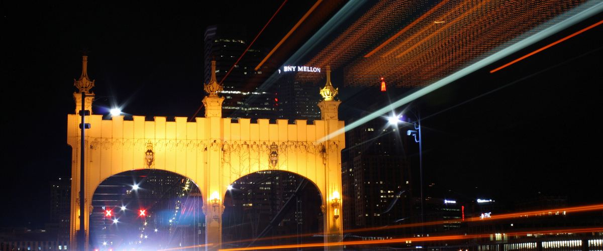 The lit-up gateway of the Smithfield Street Bridge at night with streaks of car lights and downtown towers behind it