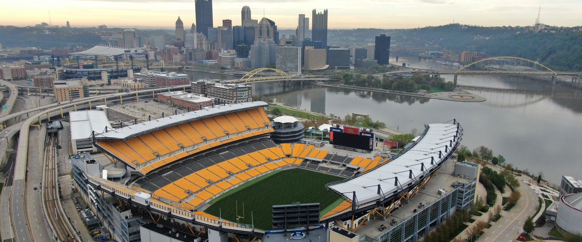 Aerial view of the North Shore football stadium with its yellow seats and empty grass field, the Allegheny River, yellow bridges and the downtown Pittsburgh skyline behind it