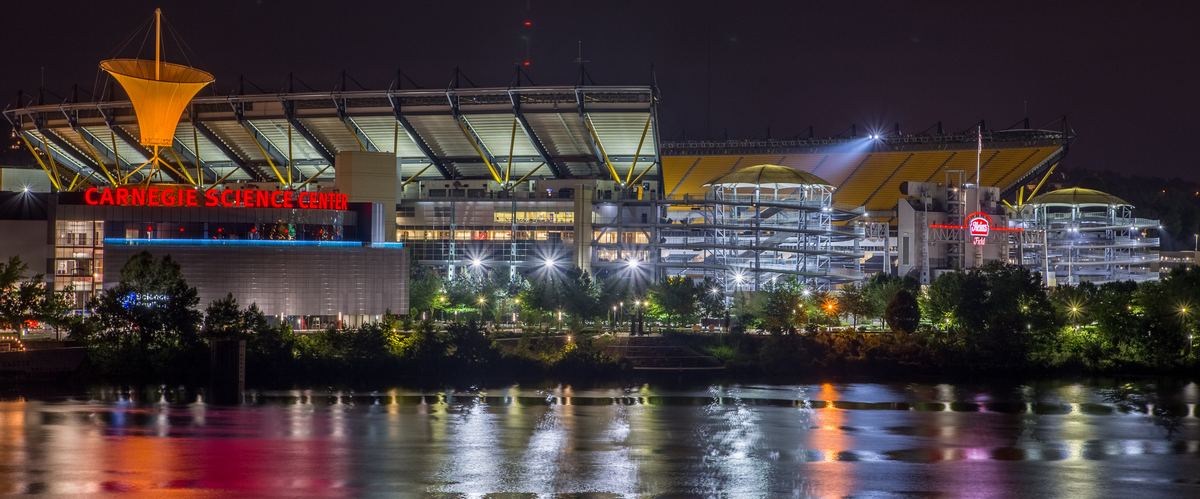 The North Shore football stadium lit up at night beside the Carnegie Science Center, with colorful lights reflected in the river